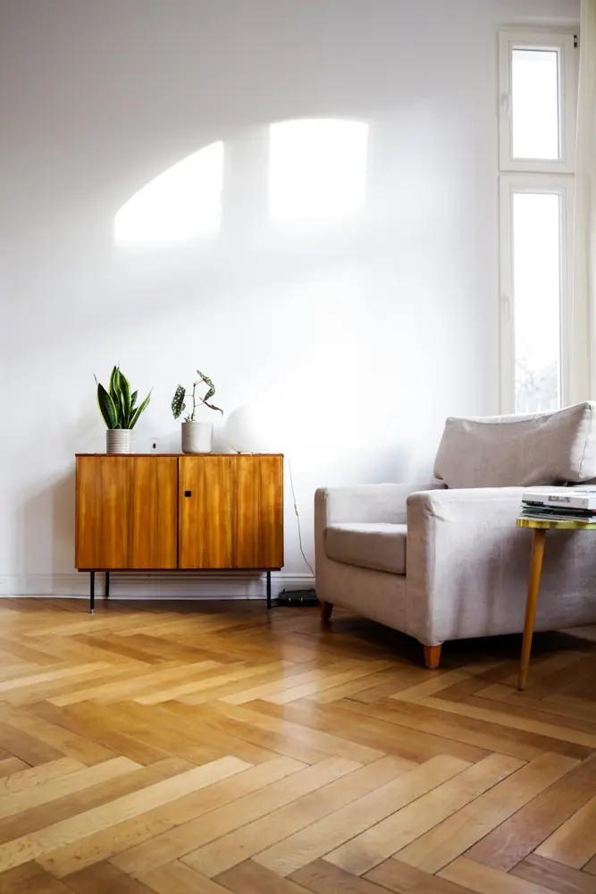 Walnut sideboard with two plants beside a pale armchair on herringbone parquet